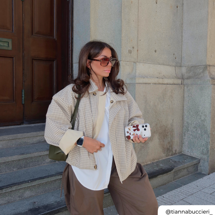 A woman stands on stone steps wearing a beige and white gingham cropped jacket over a white top and brown trousers, carrying a green shoulder bag and holding a phone in a clear phone case decorated with a red bow, cherries, a martini glass, a strawberry dipped in chocolate, and small star accents.