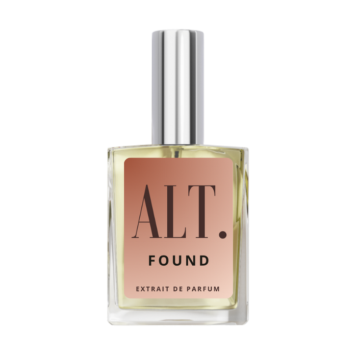 A bottle of ALT. FOUND Extrait de Parfum. Phlur Missing Person Dupe with a square clear glass body, pale yellow liquid, a peach-pink label with black text, and a reflective silver cap, centered on a white background.