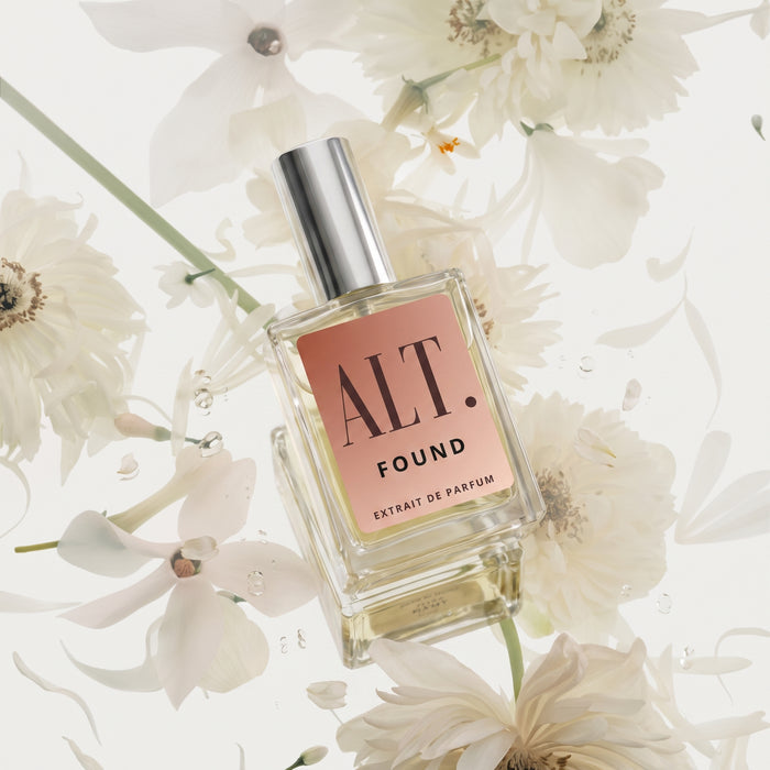 A clear glass perfume inspired by Phlur Missing Person with a silver cap and a dusty pink label reading 'ALT. FOUND EXTRAIT DE PARFUM', surrounded by white flowers on a light background.