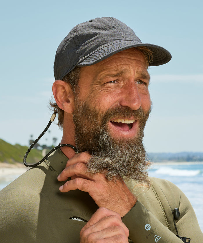 Man with a beard and cap standing by the water, wearing a wetsuit.