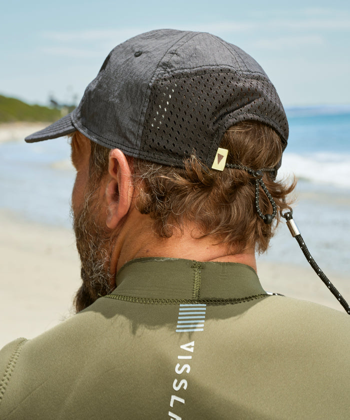 Person wearing a cap and green jacket with 'Vissla' branding on a beach.