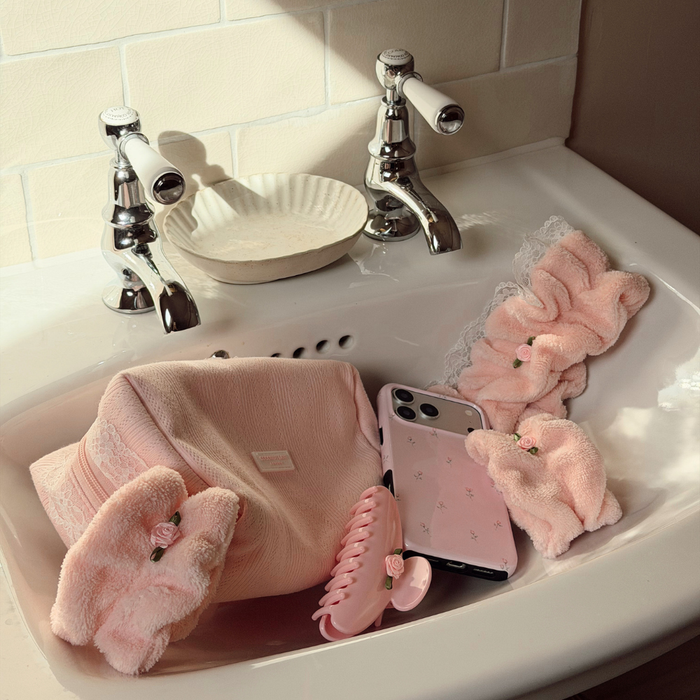 A collection of soft pink bath accessories arranged in a white sink, including plush bath towels, a claw hair clip, a phone case, and a scrunchie, with chrome faucets and a tiled wall in the background.